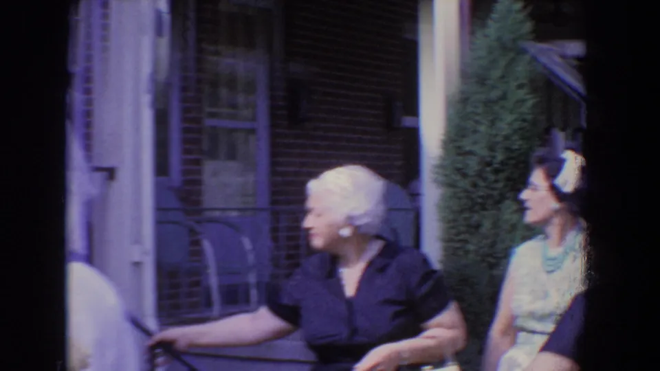 1960 Bel Air home-movie frame showing dressed-up adults near a brick porch, with an older woman in a dark dress at left.