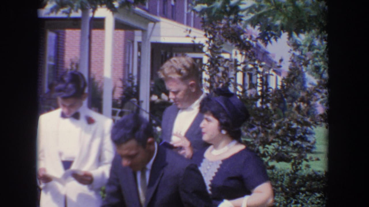 1960 Bel Air footage shows adults in formal clothing outside a residence, with a man in a white tuxedo looking down at paper.