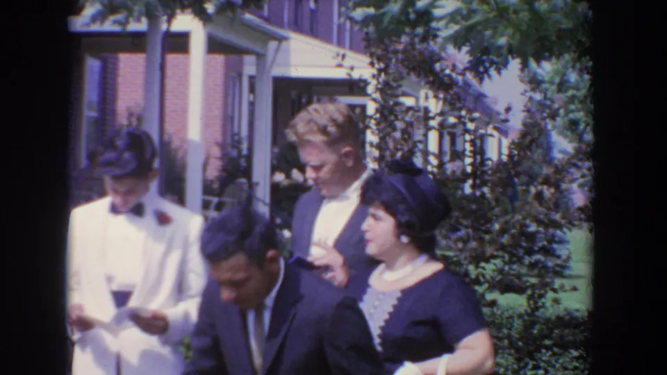 1960 Bel Air footage shows adults in formal clothing outside a residence, with a man in a white tuxedo looking down at paper.