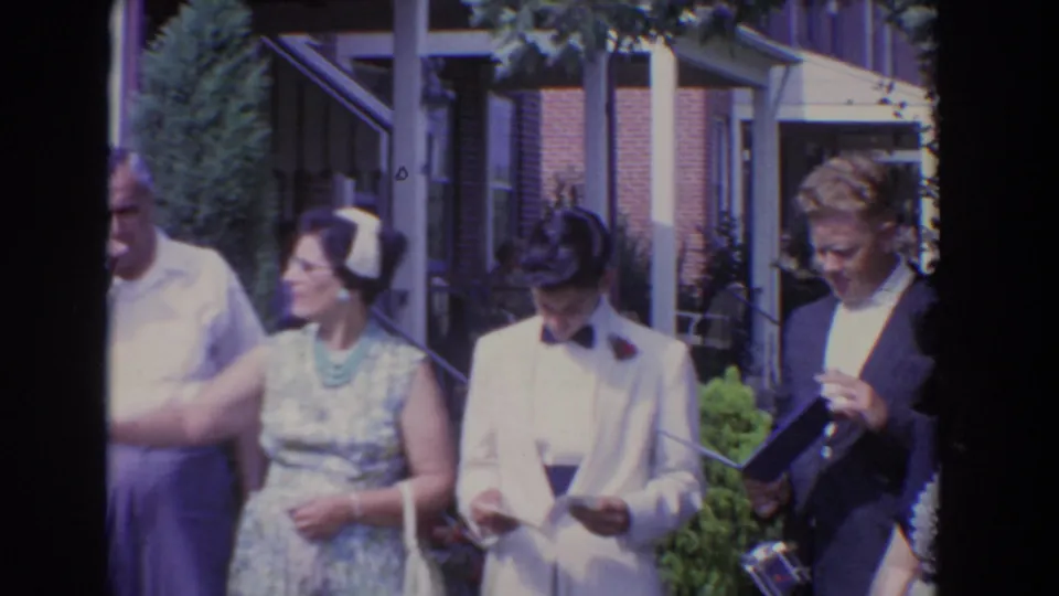 Adults in 1960 Bel Air stand close together outside a brick home, including a woman in a patterned dress and a man in a white tuxedo.