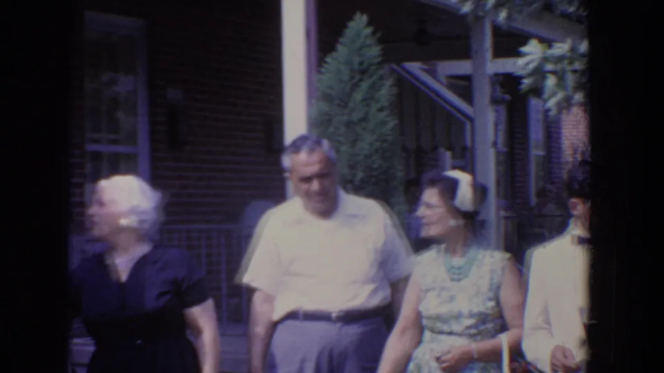1960 Bel Air home-movie frame of dressed-up adults moving past a brick residential porch in strong outdoor light.