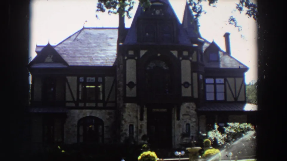 The residence fills the frame, with half-timber trim, stonework, garden plantings, and sprinkler spray visible at the lower right.