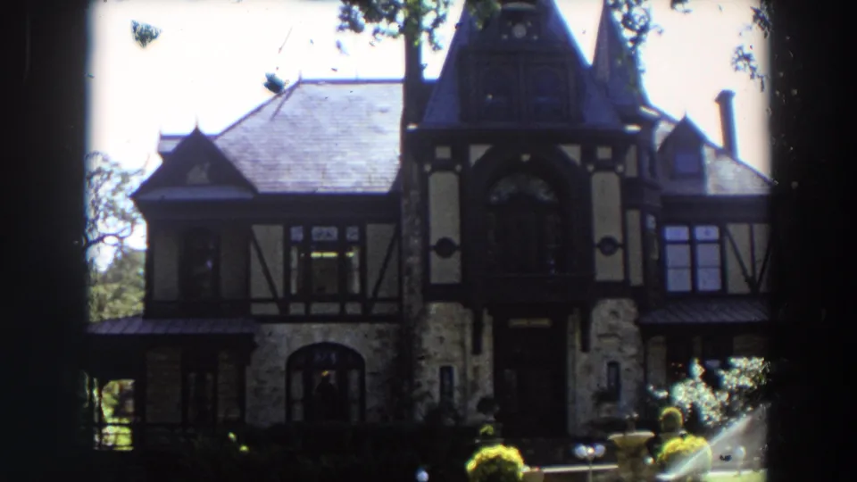 A slightly closer 8mm view shows the steep roof, dark trim, stone lower level, shaded entry, and moving sprinkler spray.