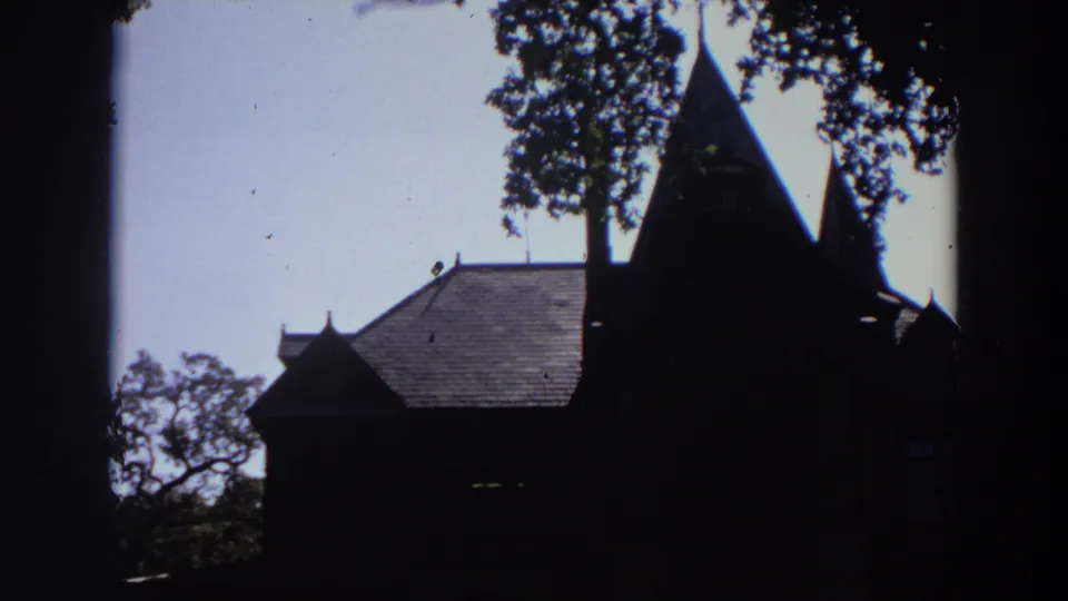 1964 California home-movie frame of a dark chapel-like building with steep spires, leafy branches, and birds in the sky.