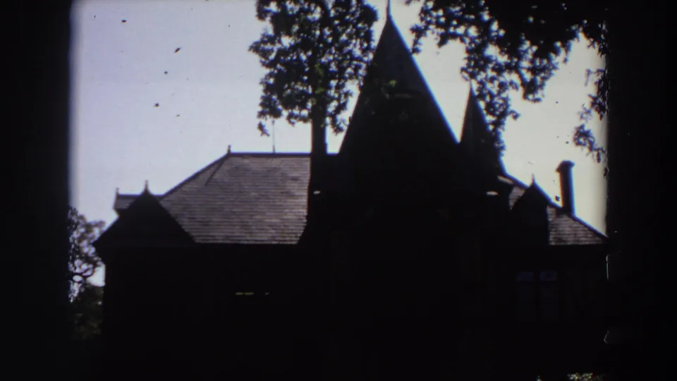 1964 California 8mm frame showing a dark, tree-framed view of a steep-roofed building near the winery grounds.