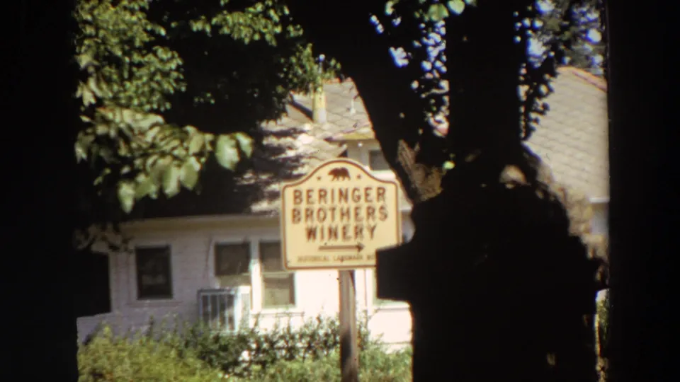1964 California 8mm frame of the Beringer Brothers Winery sign, leafy branches, tree trunks, and a pale winery building behind.