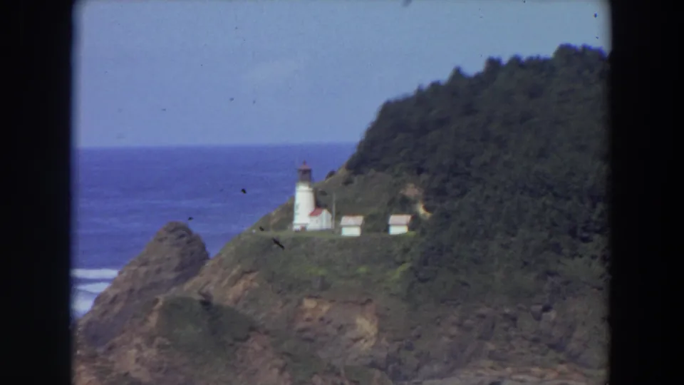 1964 California 8mm frame with a white lighthouse and small structures on a wooded coastal bluff above blue water.