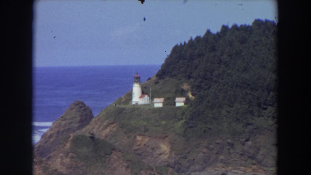White lighthouse and outbuildings on a rocky forested cliff above blue ocean.