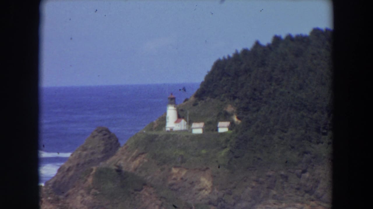 Distant lighthouse complex on a green cliff with ocean and forested hill behind.