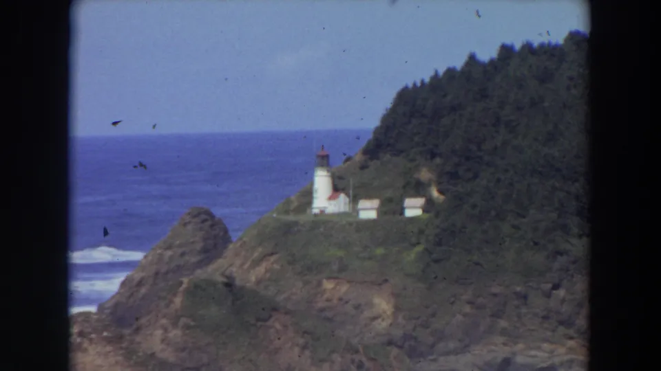 A soft 8mm frame shows the California coastal lighthouse from a distance, with surf at the rocks and a forested slope nearby.