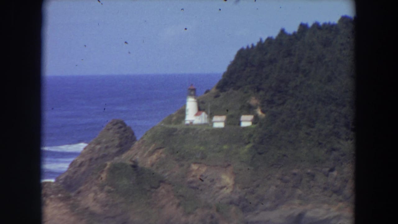 1964 California 8mm frame showing a distant white lighthouse on a green headland above the Pacific Ocean.