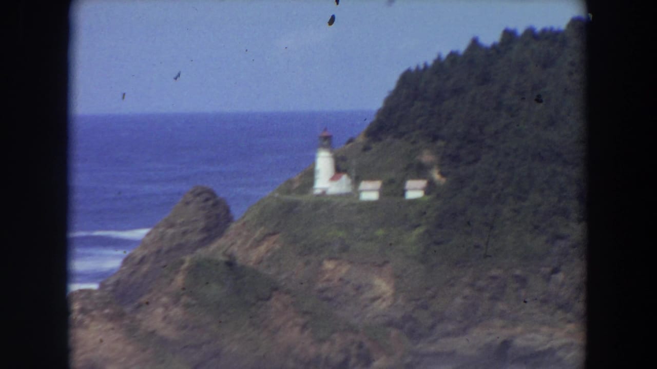 Distant lighthouse complex on a forested coastal headland beside the ocean.
