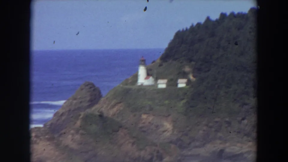 1964 California coastal view with a white lighthouse and outbuildings on a green hill over blue water.