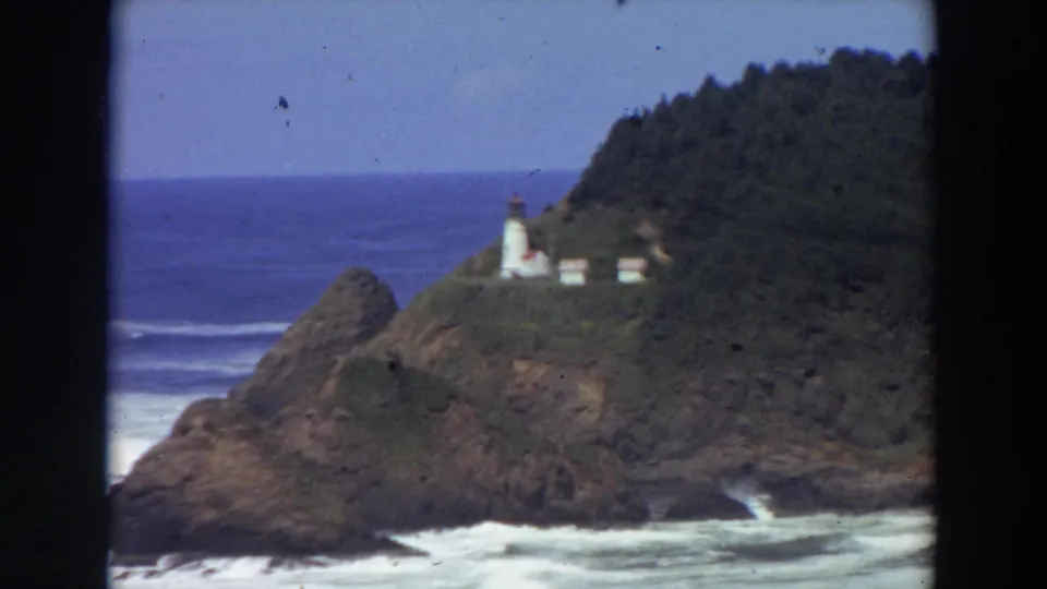 1964 California home-movie frame showing a white lighthouse and small buildings above a rocky Pacific shoreline.