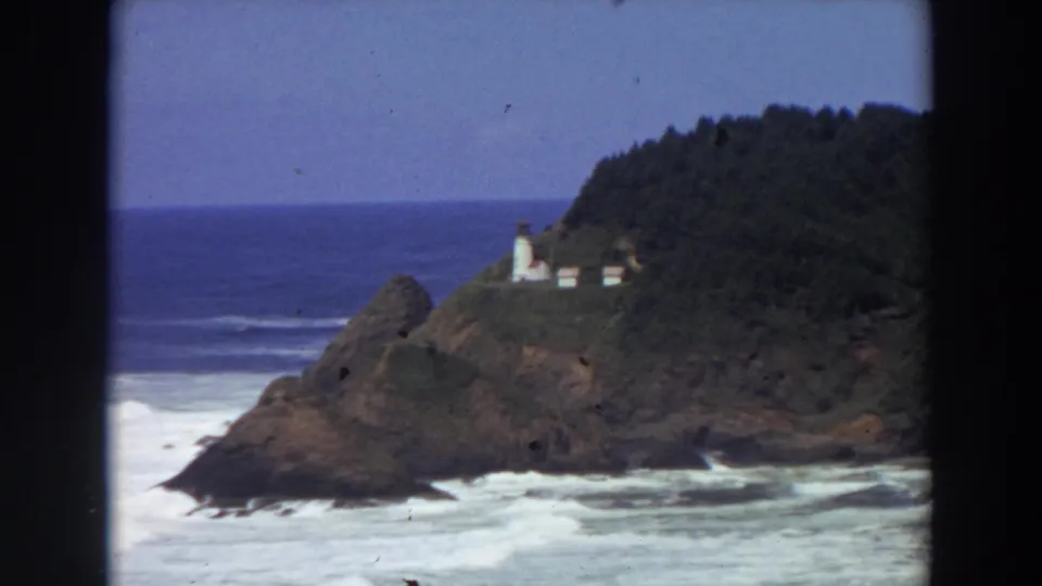 1964 California 8mm view of a white lighthouse and small buildings on a rocky Pacific headland with waves below.
