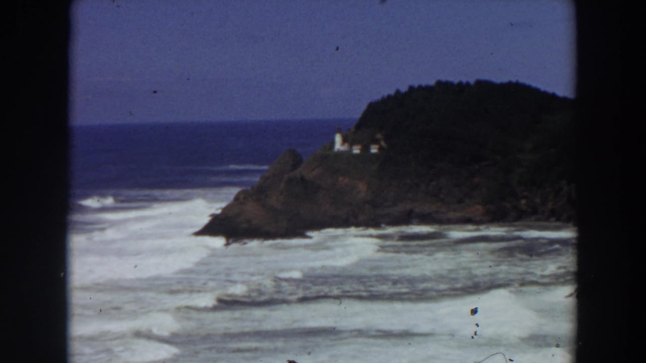 Rocky coastal point with lighthouse, trees, blue ocean, and white surf.