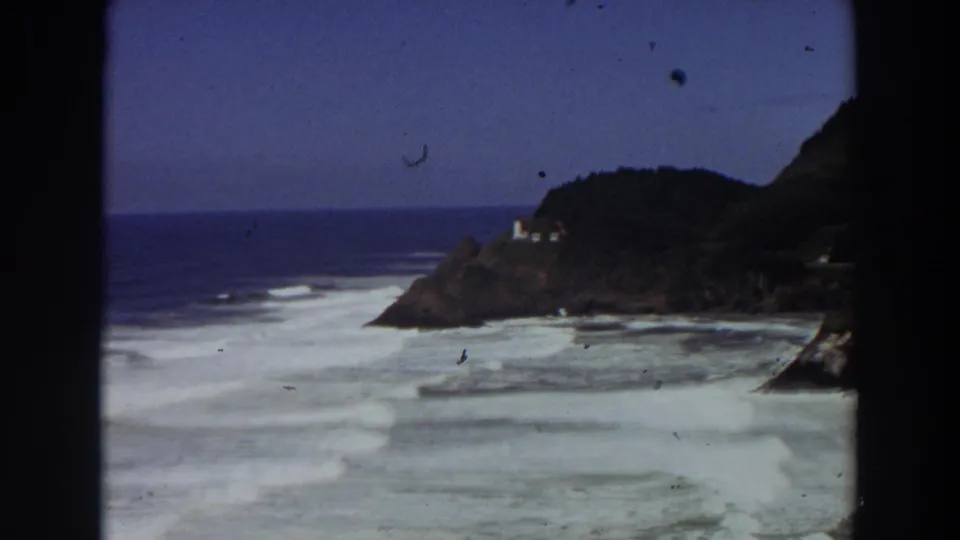 Wide 1964 California home-movie frame of rolling surf and a distant lighthouse on a dark wooded headland.