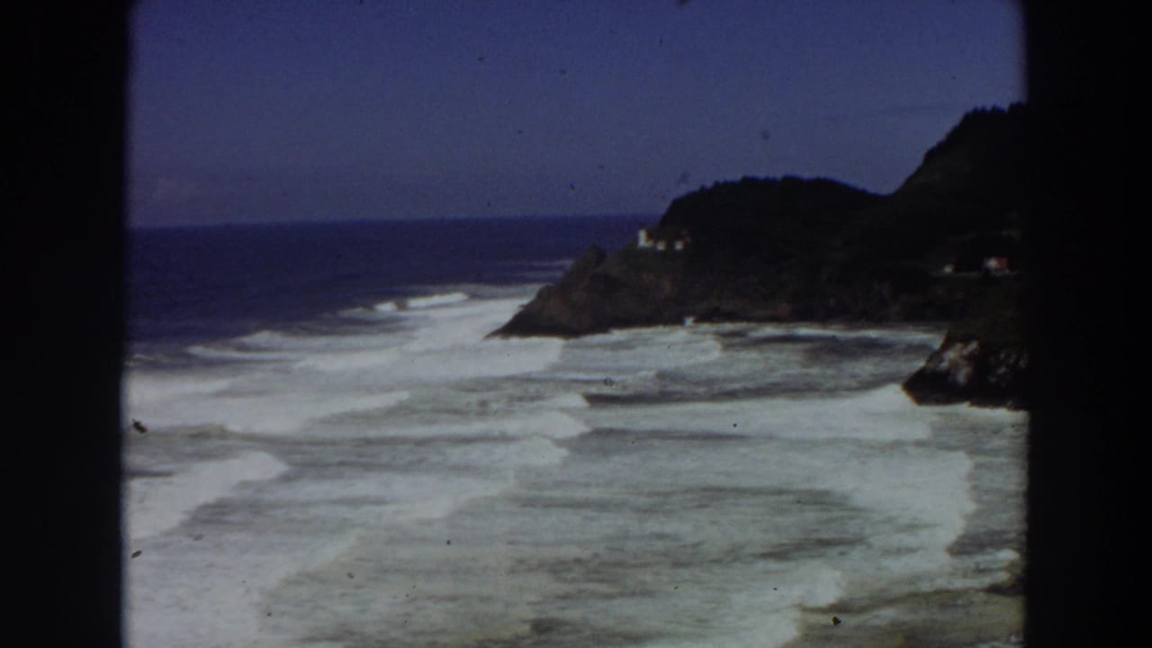 1964 8mm California coastal view with white surf, blue ocean, rocky cliff, and a small pale building near the headland.
