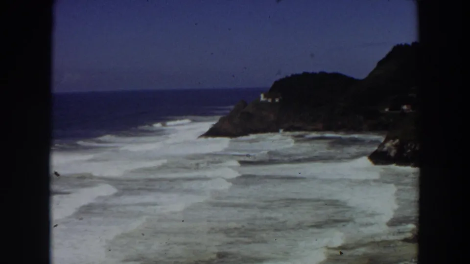 1964 8mm California coastal view with white surf, blue ocean, rocky cliff, and a small pale building near the headland.