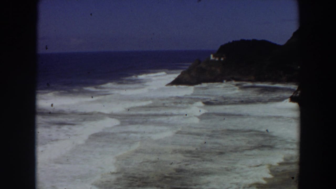 1964 California coast frame showing white breaking waves, reef-like shallows, blue horizon, and a small structure on the rocky point.