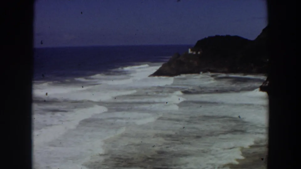 1964 California coast frame showing white breaking waves, reef-like shallows, blue horizon, and a small structure on the rocky point.