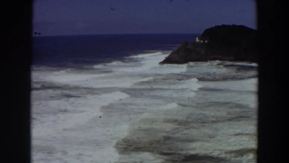Vintage 8mm California shoreline view with foamy surf in the foreground and a dark cliff carrying a small pale building beyond.