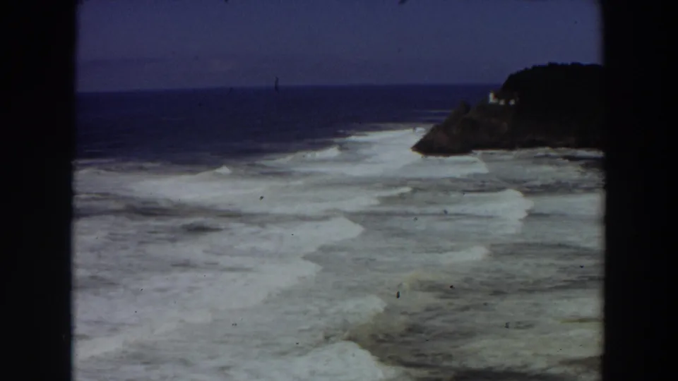 1964 8mm frame of ocean waves and a rocky California shoreline, with dark film or window edges framing the view.