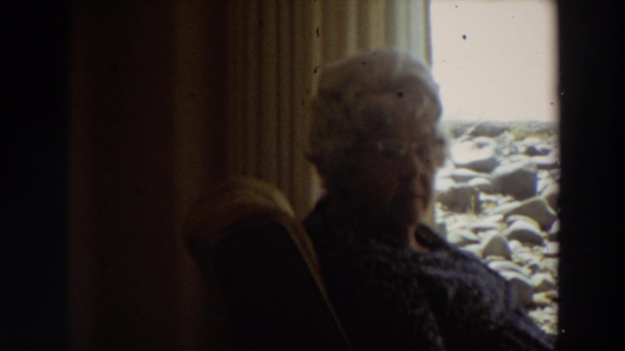 Older woman with white hair and glasses sits in a dim California interior beside a window looking toward rocks and surf.