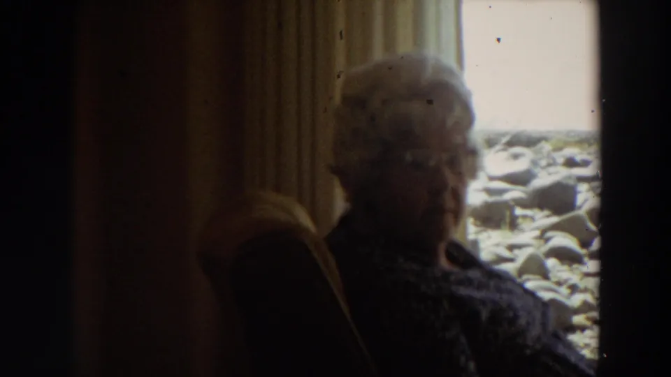 Older woman with white hair and glasses sits in a dim California interior beside a window looking toward rocks and surf.