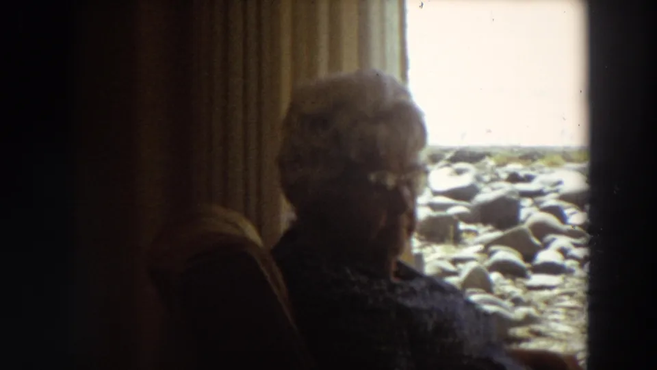 1964 home-movie frame of an older woman in a dim room, seated near curtains with a rocky California shoreline beyond the window.