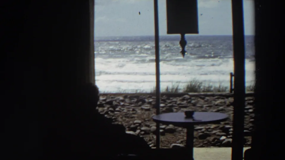1964 California home-movie frame with a dark seated silhouette, round table, window frame, rocky shore, and churning ocean.