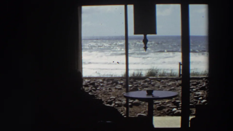 1964 California 8mm frame of a seated silhouette inside a dim room, facing a bright window with rocky beach and surf beyond.