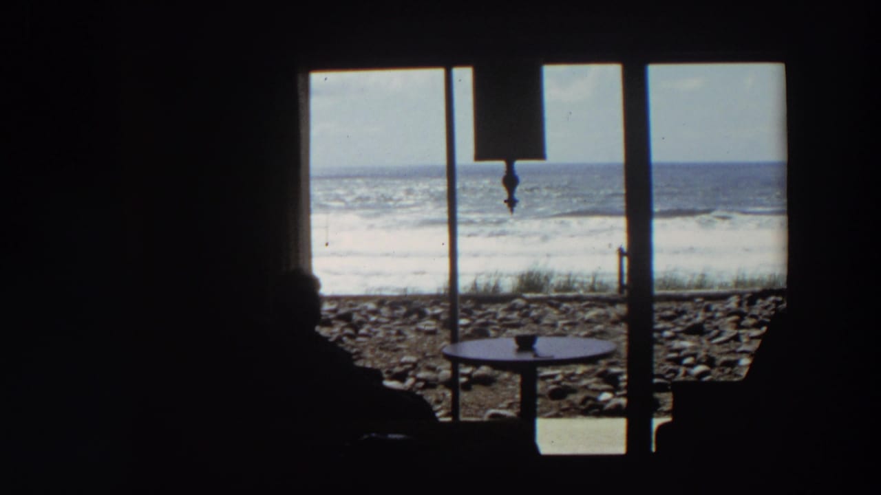 California coastal home-movie frame showing a person in silhouette near a window as waves break beyond a rocky beach.
