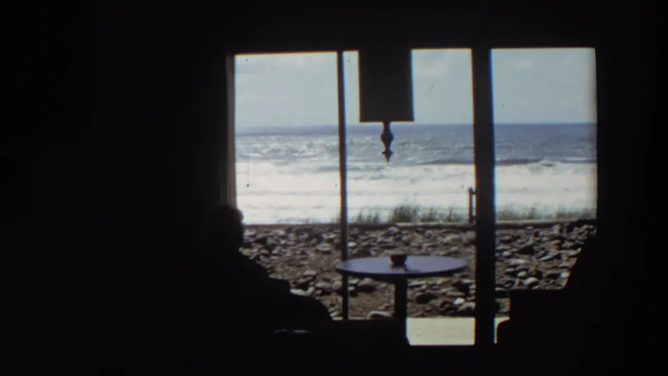 California coastal home-movie frame showing a person in silhouette near a window as waves break beyond a rocky beach.