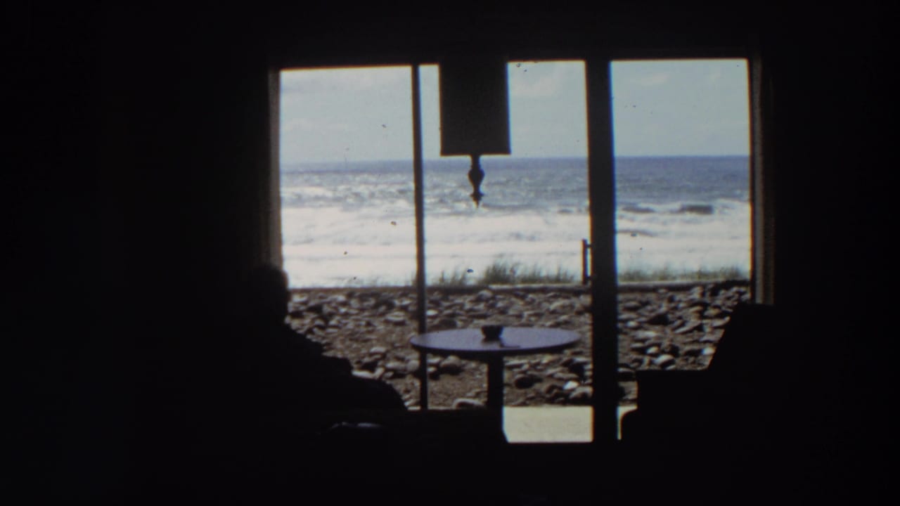 1964 California home-movie frame with dark interior silhouettes, a small table, sliding glass doors, stones, surf, and ocean horizon visible outside.