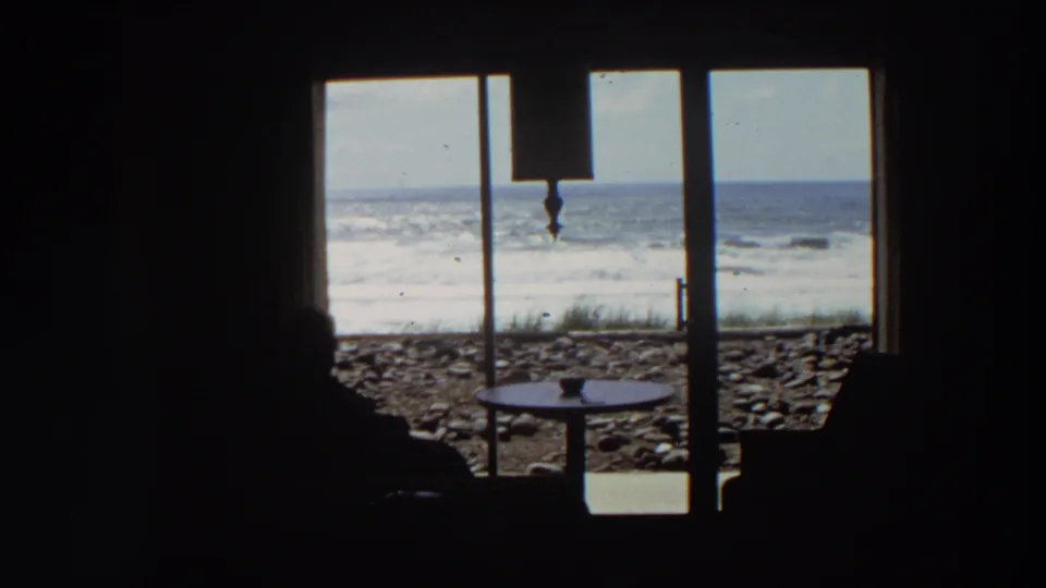 1964 California home-movie frame with dark interior silhouettes, a small table, sliding glass doors, stones, surf, and ocean horizon visible outside.