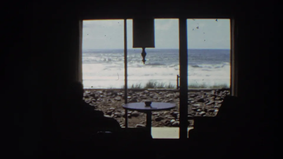 1964 California 8mm view from inside a coastal room, with silhouetted seated figures, a round table, glass doors, rocky beach, and surf.