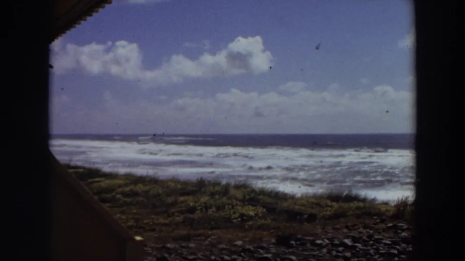 1964 California coastal home-movie frame looking out toward surf, rocky shoreline, low coastal plants, ocean horizon, and a partly cloudy sky.
