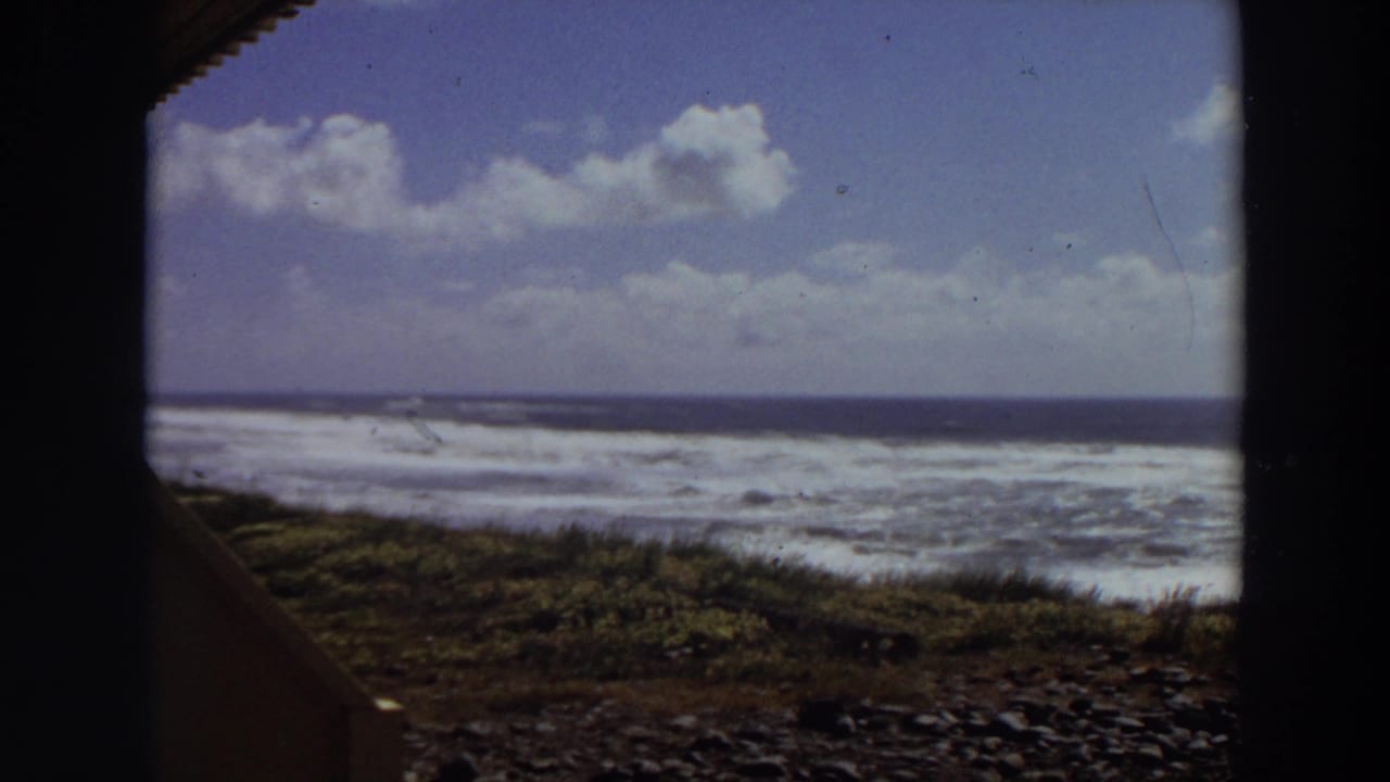 1964 California 8mm frame showing whitewater waves beyond a rocky beach and low coastal vegetation, seen through dark frame edges.