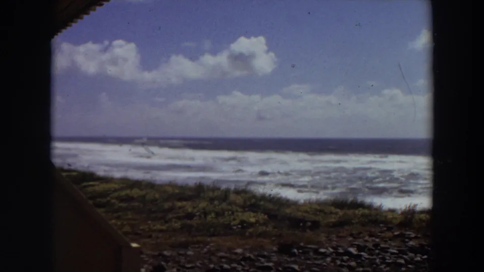 1964 California 8mm frame showing whitewater waves beyond a rocky beach and low coastal vegetation, seen through dark frame edges.