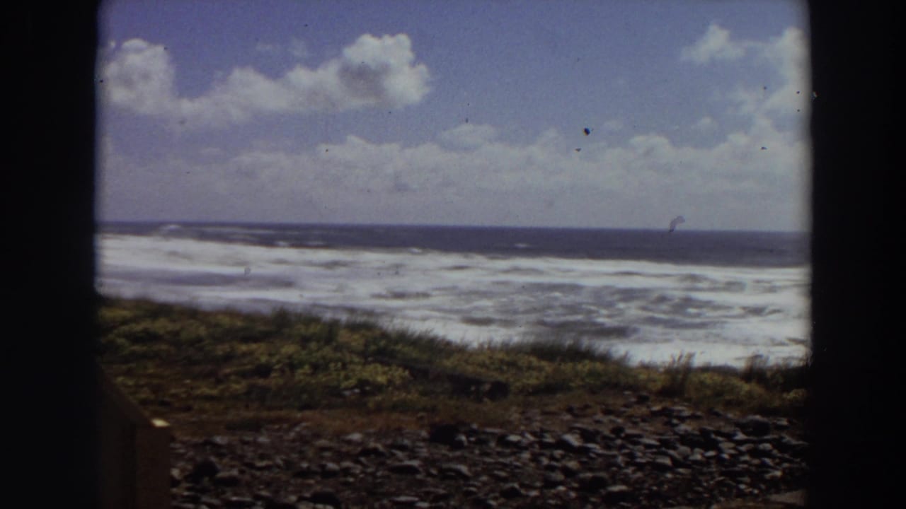 1964 California coastal home-movie frame with turbulent surf, dark ocean horizon, grassy slope, and rounded 8mm frame edges.