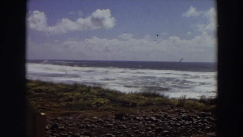 1964 California coastal home-movie frame with turbulent surf, dark ocean horizon, grassy slope, and rounded 8mm frame edges.