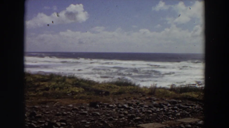 1964 California 8mm still of a rocky vegetated beach with white surf, offshore horizon, and bright clouds in the sky.