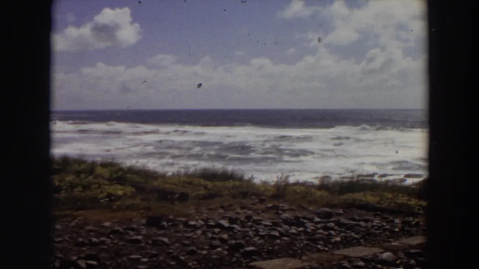 1964 California coastal frame showing foamy surf, pebbled foreground, low vegetation, and a cloudy blue sky through an 8mm frame.