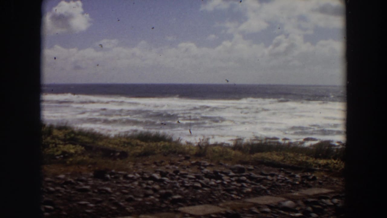 1964 California 8mm frame showing turbulent ocean waves, rocky foreground, low grass, and cloudy sky within dark film-gate edges.