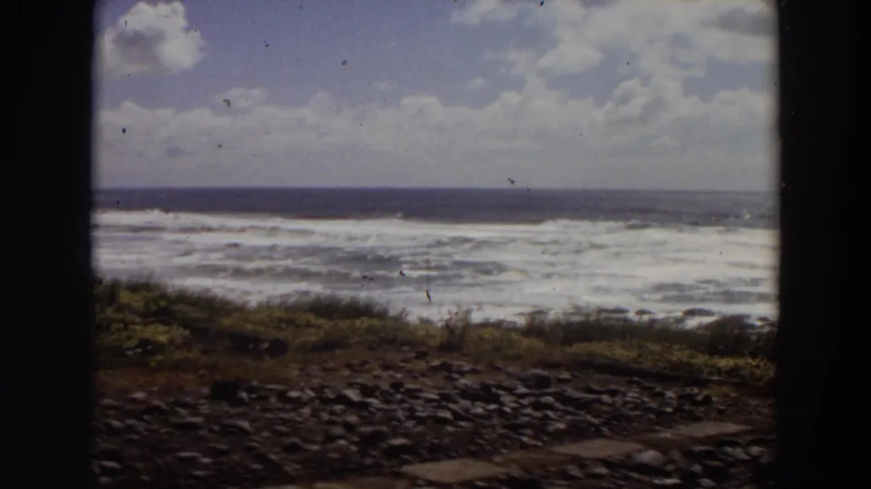 1964 California 8mm frame showing turbulent ocean waves, rocky foreground, low grass, and cloudy sky within dark film-gate edges.