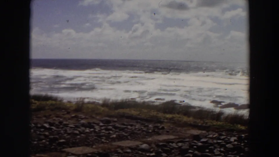 Vintage 8mm frame from a 1964 California coast scene, showing rocky ground, low vegetation, bright surf, and a distant horizon.