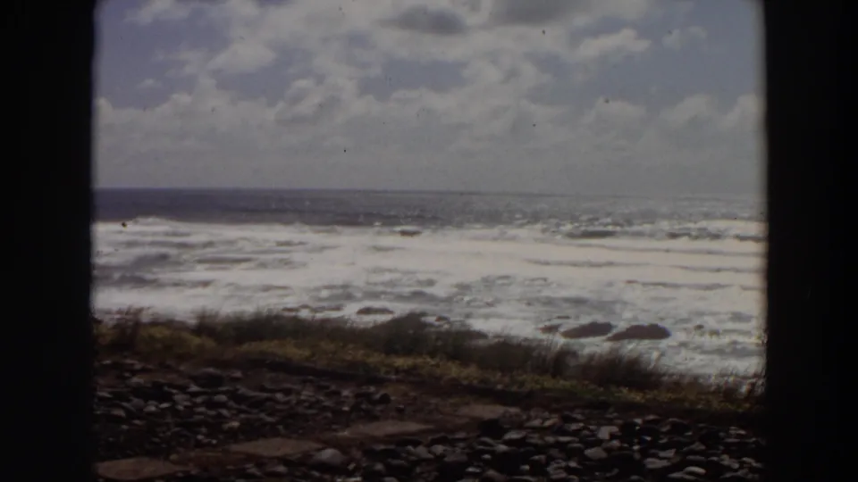 1964 California home-movie still showing a wide ocean horizon, white surf, rocky shore, coastal grass, and aged 8mm scan marks.