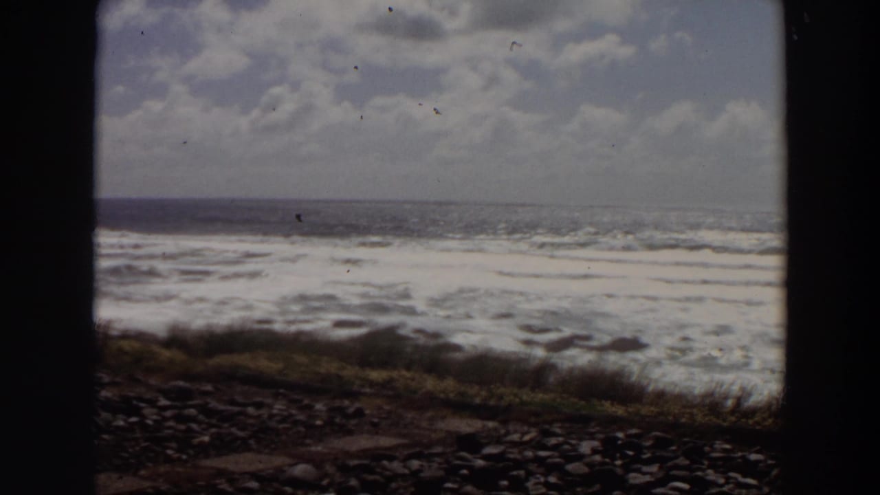 1964 California 8mm frame of foamy surf rolling toward a rocky grassy shore beneath a cloudy sky.