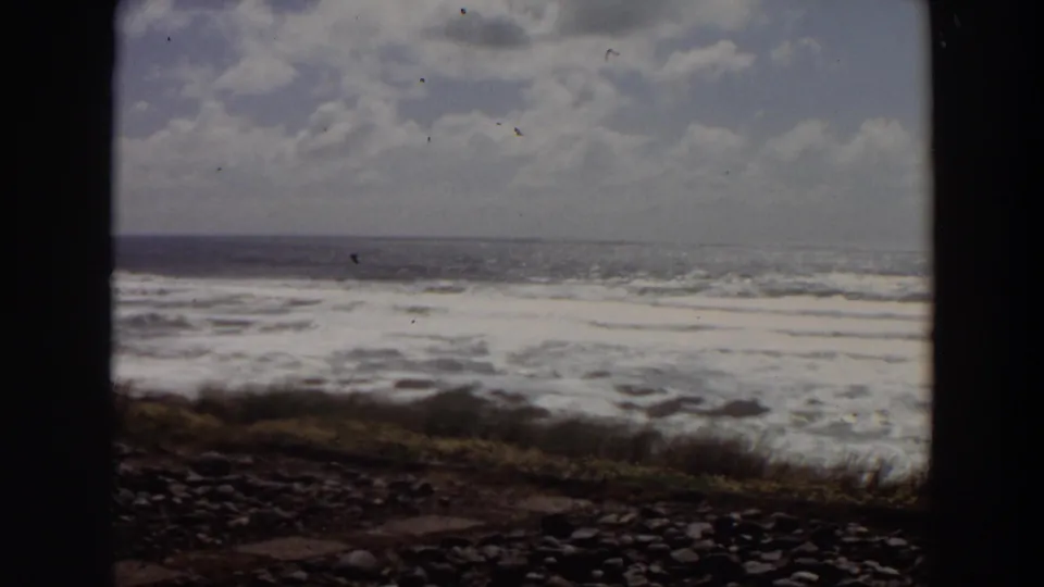 1964 California 8mm frame of foamy surf rolling toward a rocky grassy shore beneath a cloudy sky.